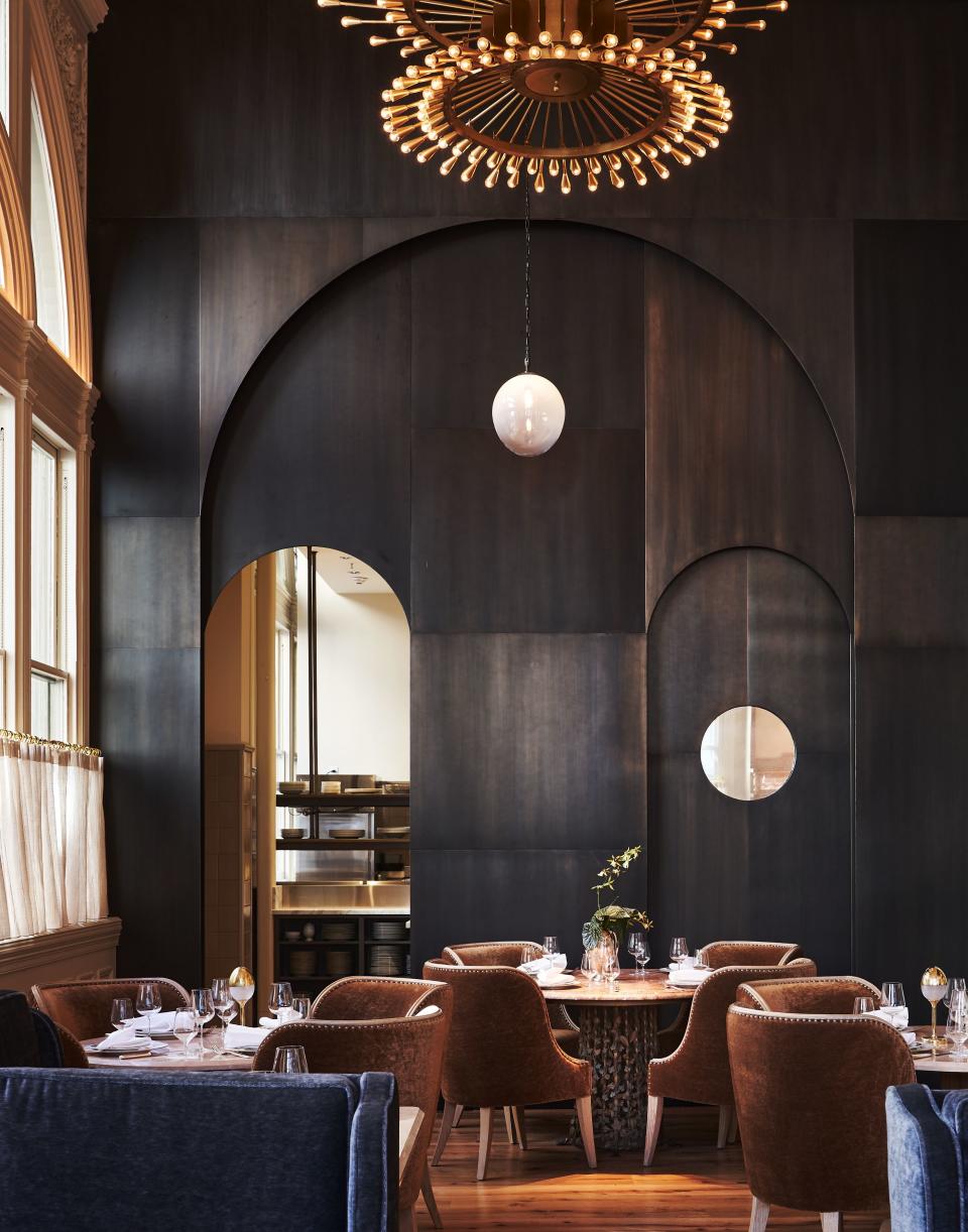 The back dining room with a view into the kitchen at Veronika, featuring blackened brass doorways and brown mohair, arched-back chairs. An RW Guild Globe light hangs in the background.