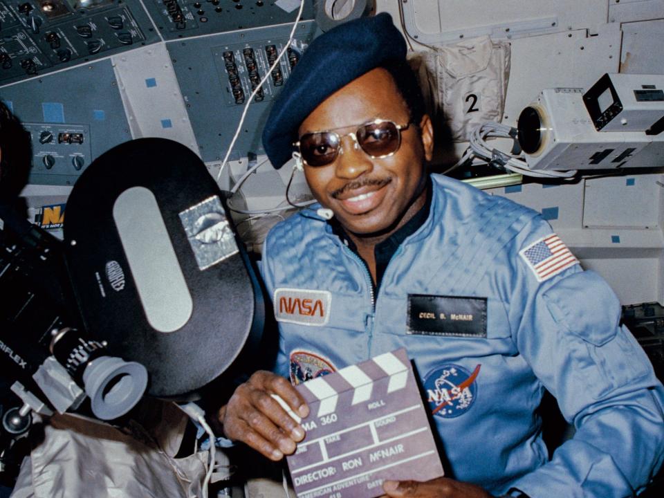 Astronaut Ron McNair wears sunglasses and a beret and holds a flim slate on a spaceshuttle