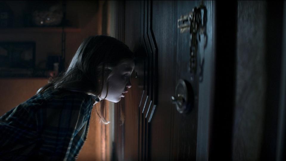 A little girl looks through a heavily locked apartment door's peephole in Evil Dead Rise.