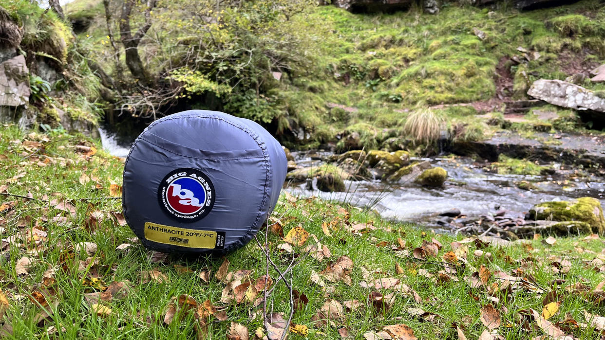  A sleeping bag in its sack, lying on grass in front of a small stream. 