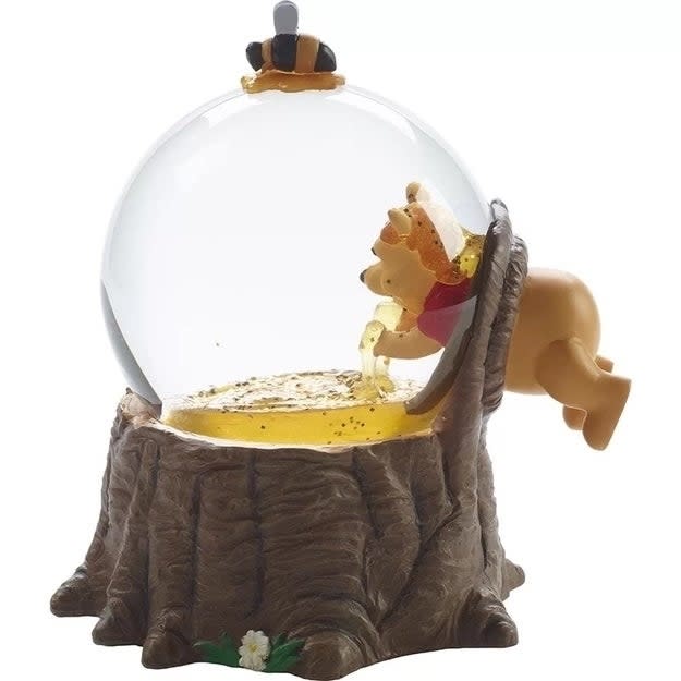 Winnie the Pooh toy figure inside a glass globe, leaning into a tree stump and reaching for honey. Top of the globe has a bee decoration