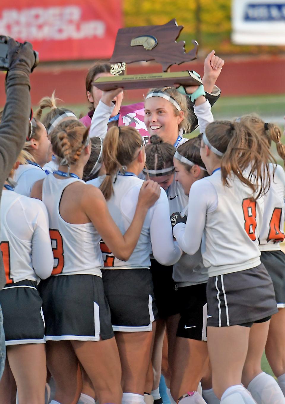 Uxbridge co-captain Tessa Johnston is surrounded by celebrating teammates as she holds the Division 4 state field hockey championship trophy.