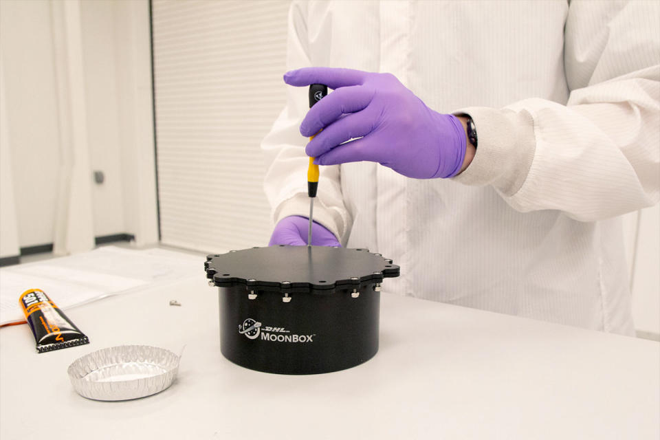 a technician in a white lab coat wearing purple gloves tightens a screw on a small black capsule.