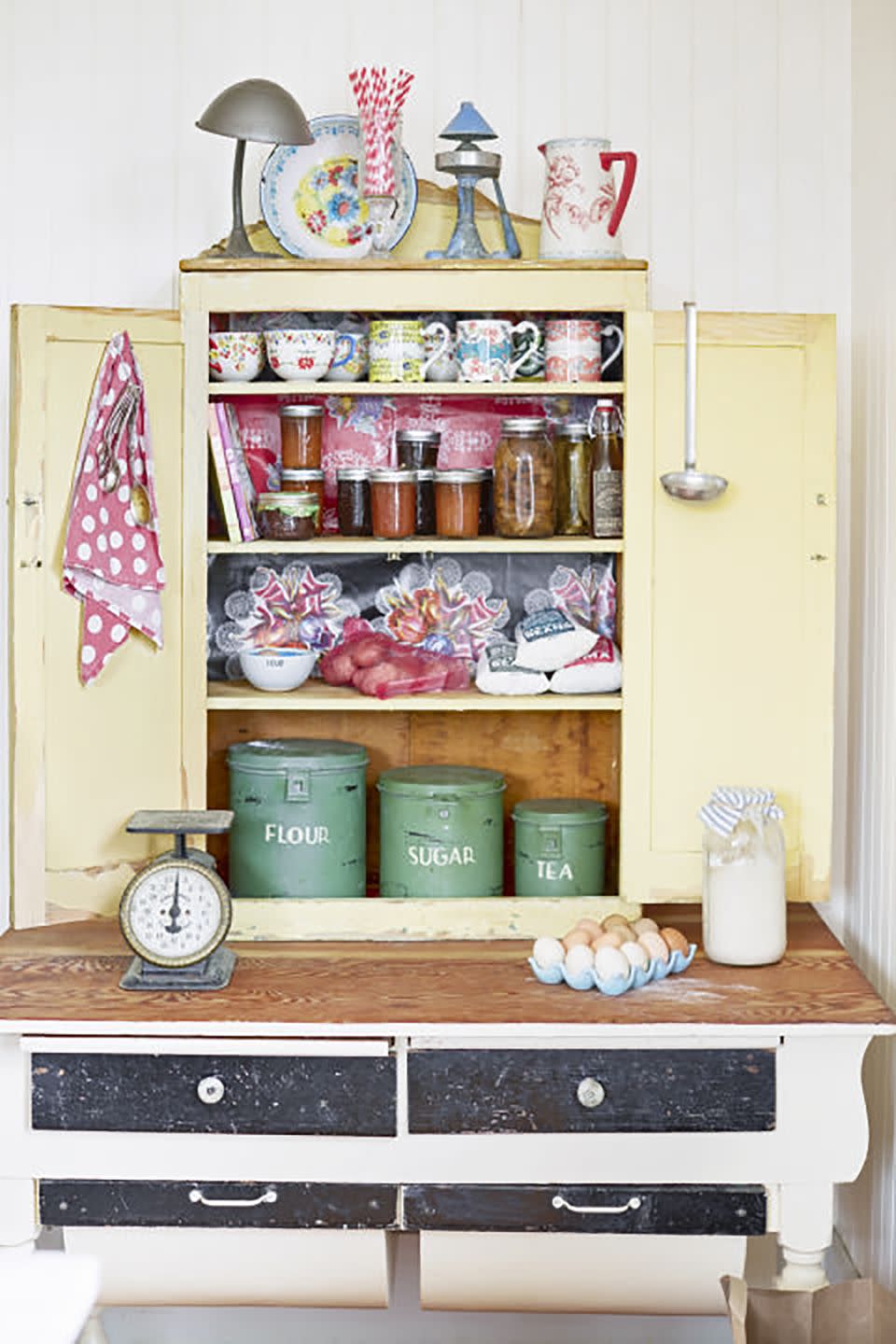 an old yellow shelf rests on top of a worktable and is filled with baking supplies and coffee mugs and jars of jams