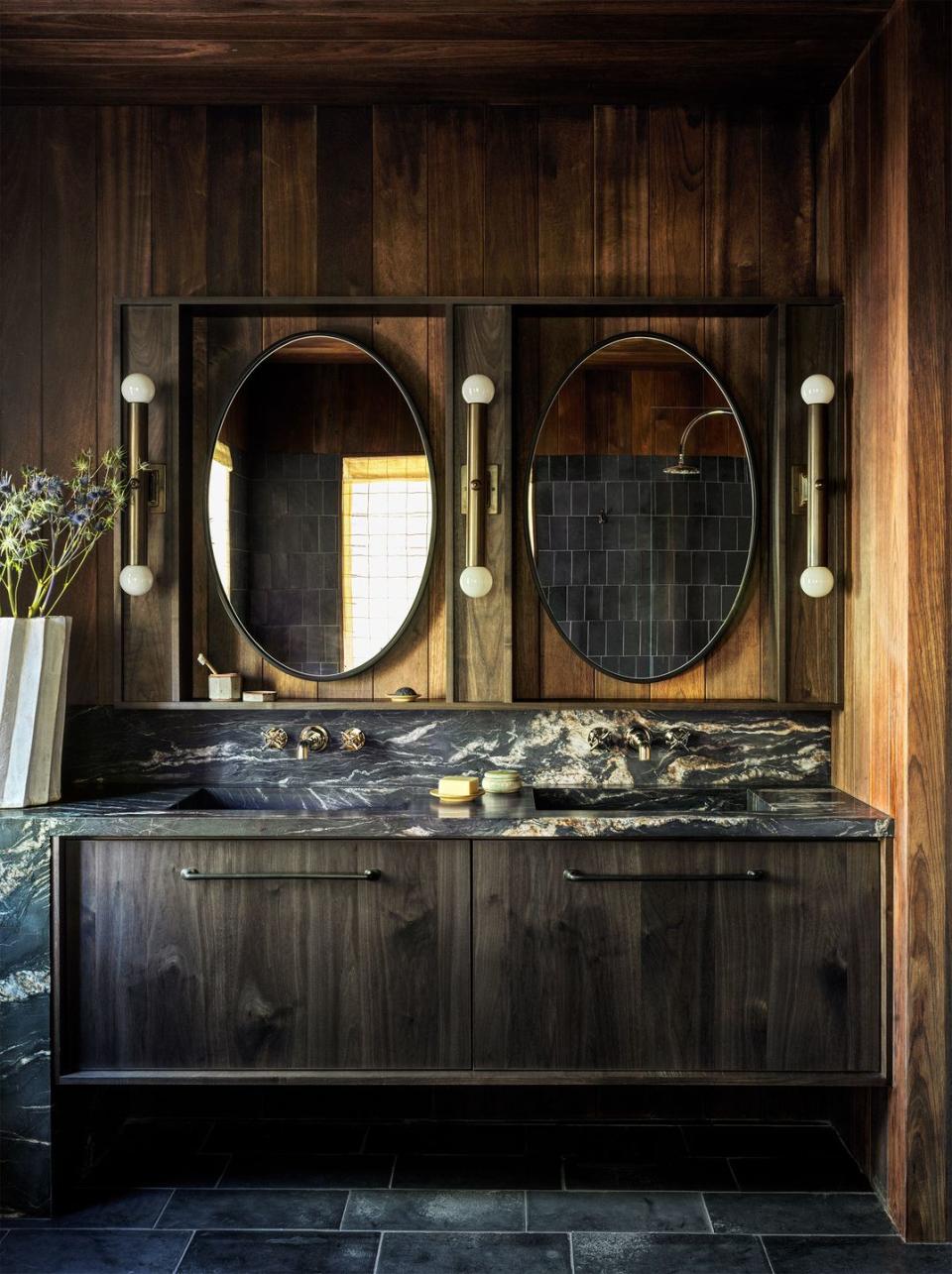 a bathroom has twin rectangular sinks in dark marble with iron walnut cabinets below, oval mirrors above each and three vertical sconces with small globes at top and bottom, spruce paneled walls, and black limestone floor