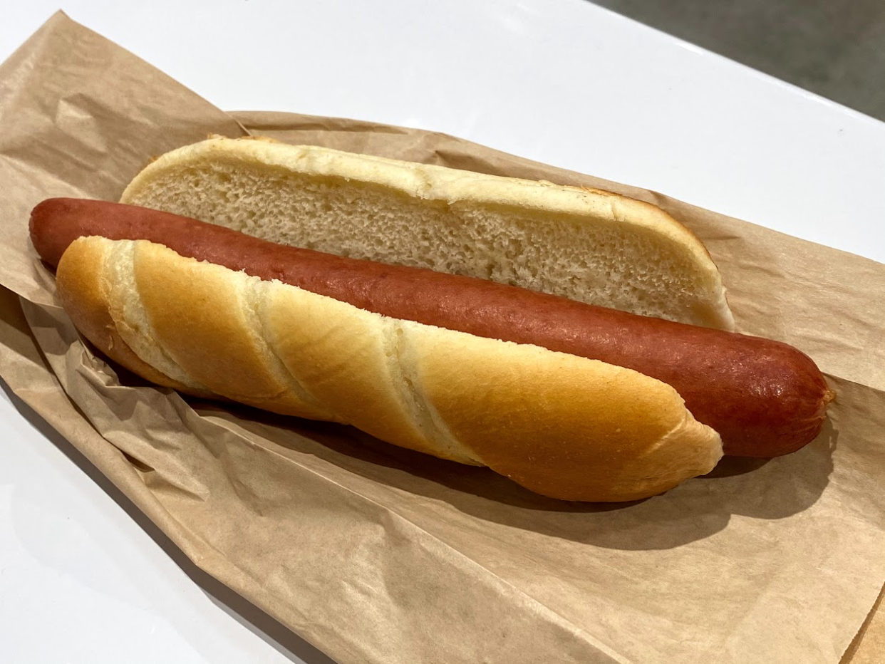 Costco hot dog, plain, on a brown paper bag on a white table, grey concrete floor in the top right background