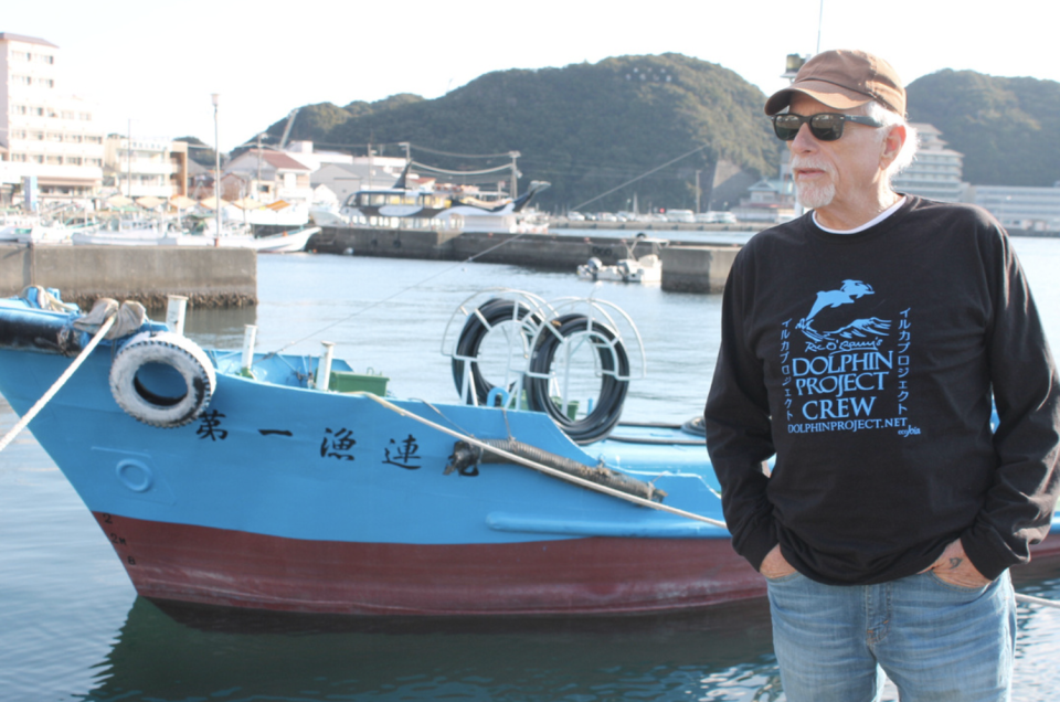 Ric O'Barry wears jeans and a Dolphin Project wind-cheater. The ocean and a boat with Japanese characters on it is in the background. 