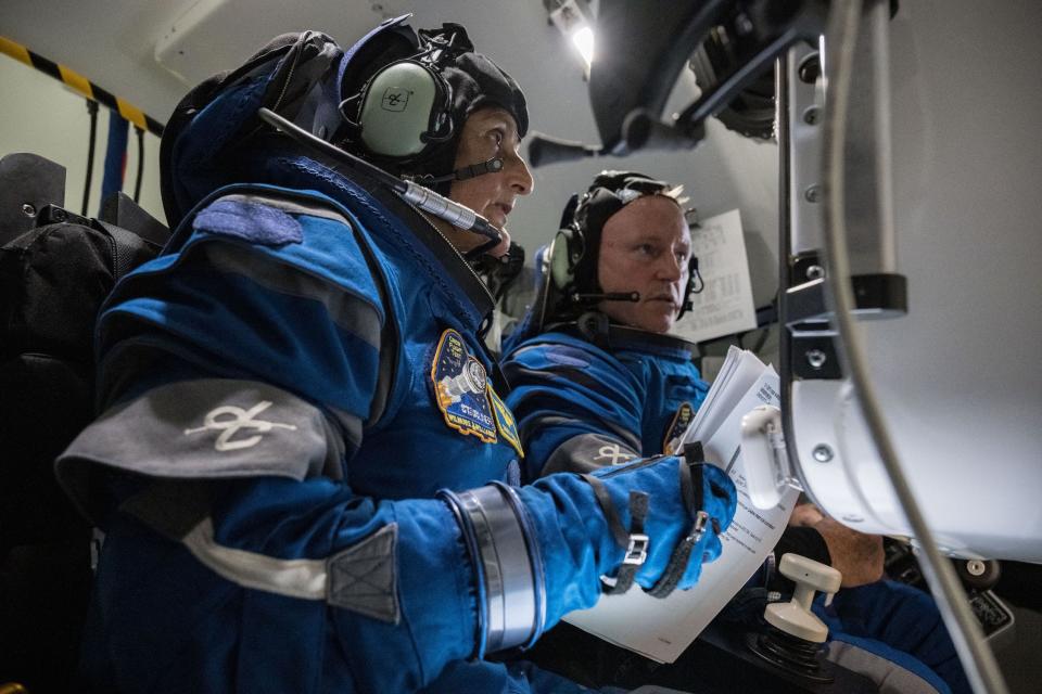 doi astronauți în costume spațiale albastre în interiorul unei nave spațiale ținând hârtii uitându-se la un tablou de bord