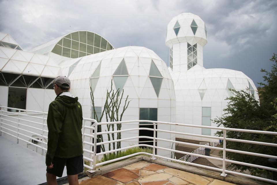 In this July 31, 2015 photo, a tourist walks to the main entrance of the Biosphere 2 facility while on a walking tour in Oracle, Ariz. University of Arizona officials say that 25 years after that New Age-style experiment in the Arizona desert, the glass-covered greenhouse thrives as a singular site for researchers from around the world studying everything from the effects of the ocean’s acidification on coral to ways of ensuring food security. (AP Photo/Ross D. Franklin)