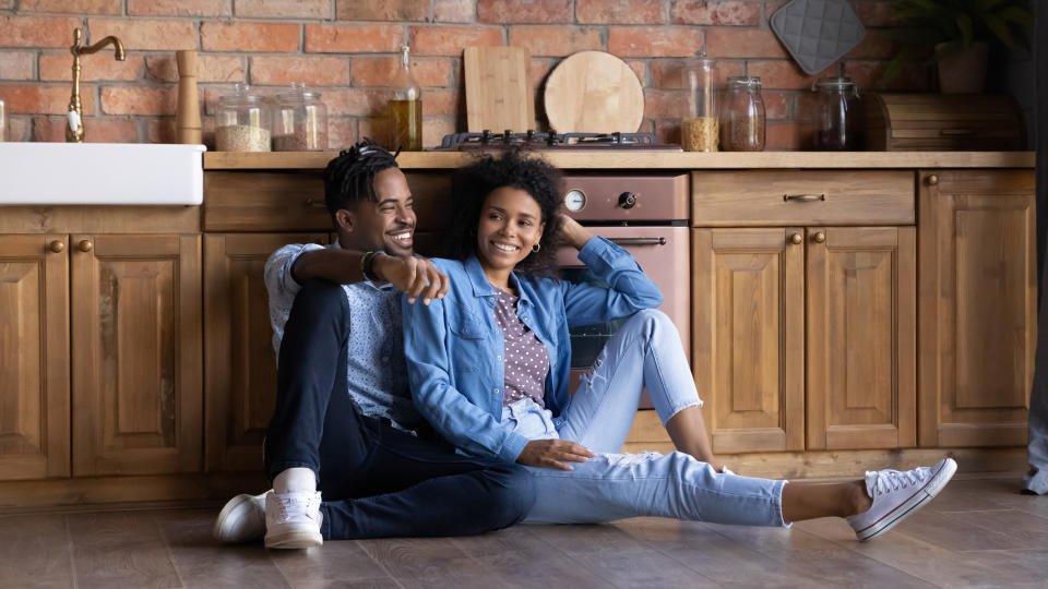 A couple, smiling and casually dressed, sit on a kitchen floor against wooden cabinets. They appear relaxed and happy