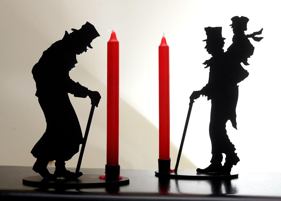 Metal candle holders with red candles and likenesses of Ebenezer Scrooge and Bob Cratchit  carrying Tiny Tim sit on a desk.