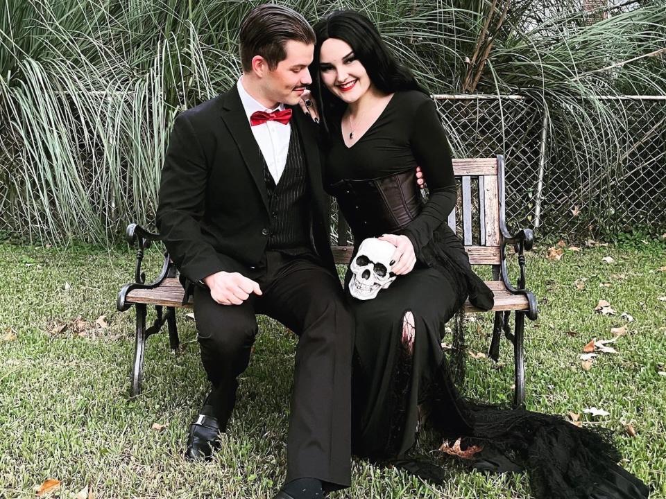 A couple dressed as Gomez and Morticia Addams sit on a bench.