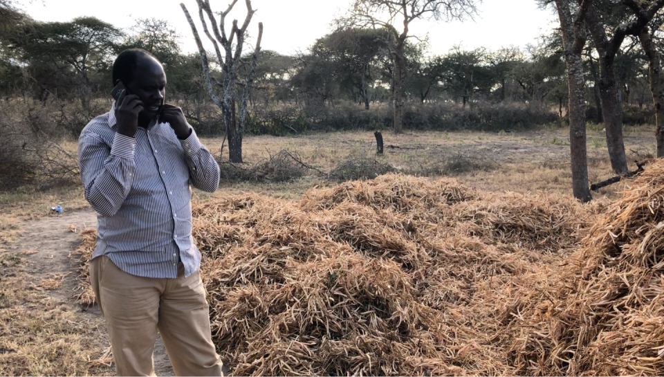 A man in a button-down shirt and trousers stands in a field with a phone held to his ear.