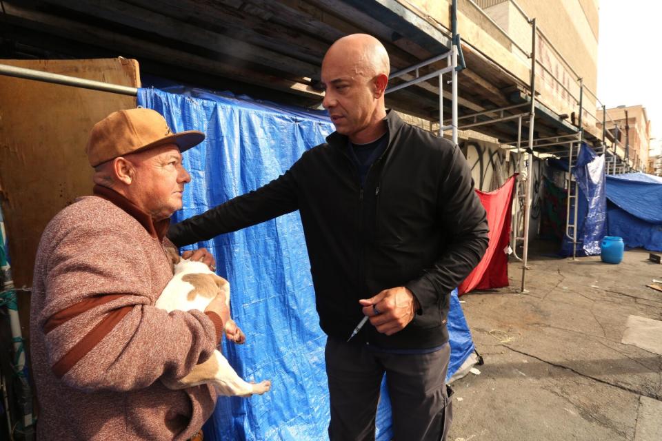 A man in a ball cap and jacket holds a dog while another man holding a syringe speaks to him.