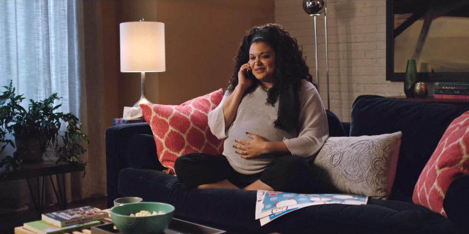 Michelle Buteau pregnant with long curly hair sits on a couch, smiling while talking on the phone, with books, a bowl of popcorn, and a children's book around her