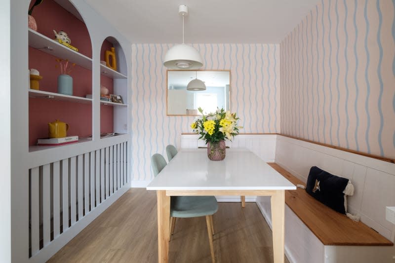 pink wallpaper with teal wavy stripe, banquet built in bench seating at white dining table with wood legs, hard wood floors, mirror above table, yellow flowers in center, light blue built in cabinets with arched top detail and pink backing, round white lighting fixture and yellow pottery accents