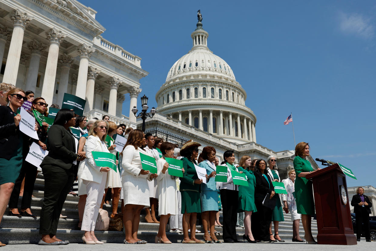 Nancy Pelosi, in a green suit, with rows of women dressed in green or white on the steps of the U.S. Capitol behind her displaying green cards with white lettering saying: Protect Women's Reproductive Freedom.