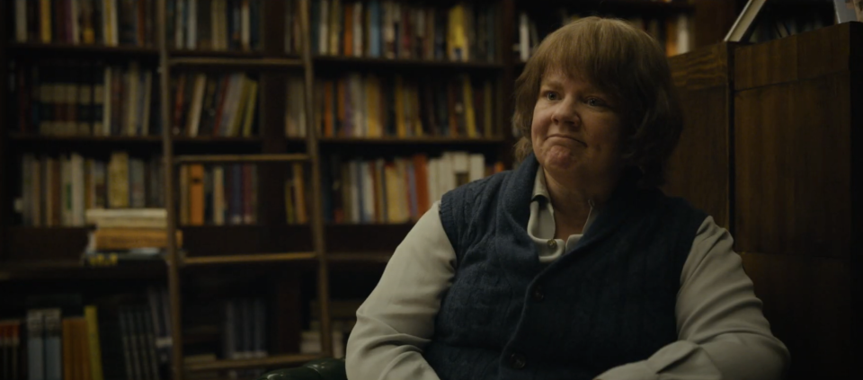 Melissa McCarthy in a library, wearing a vest and shirt, sitting and looking contemplative