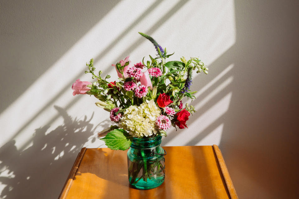 A vibrant floral arrangement in a green glass jar with various flowers including pink, red, and white blooms, placed on a wooden table