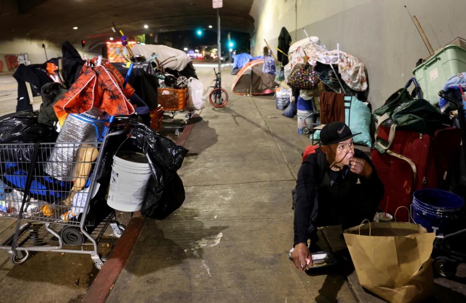 A man sits on a sidewalk with a bag next to a shopping cart