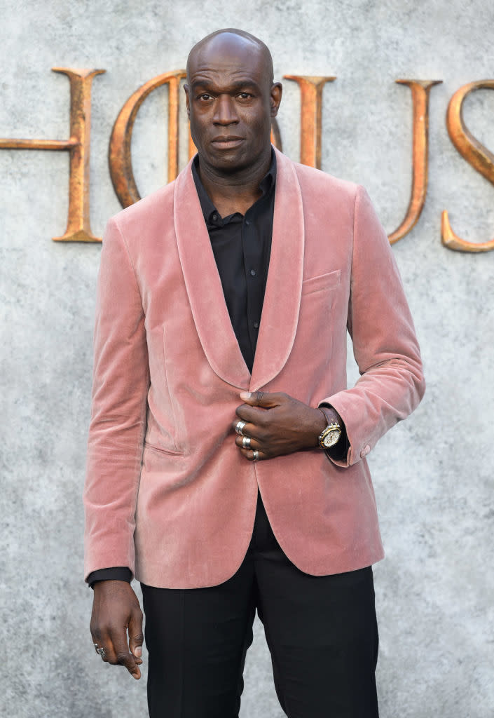 Man in a velvet blazer and dark shirt stands in front of a backdrop with large, gold text partially visible