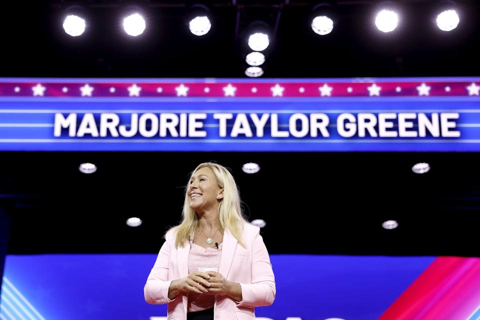 A blond woman in a pink jacket stands in front of many lights and a marquee that says 'Marjorie Taylor Greene'