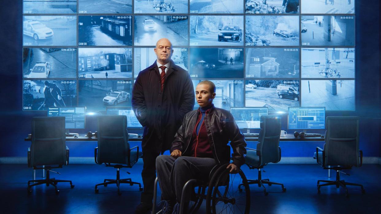  Ross Kemp as Tony stands beside Beth Alsbury in a wheelchair as Hannah in front of a bank of CCTV screens in Blindspot. 