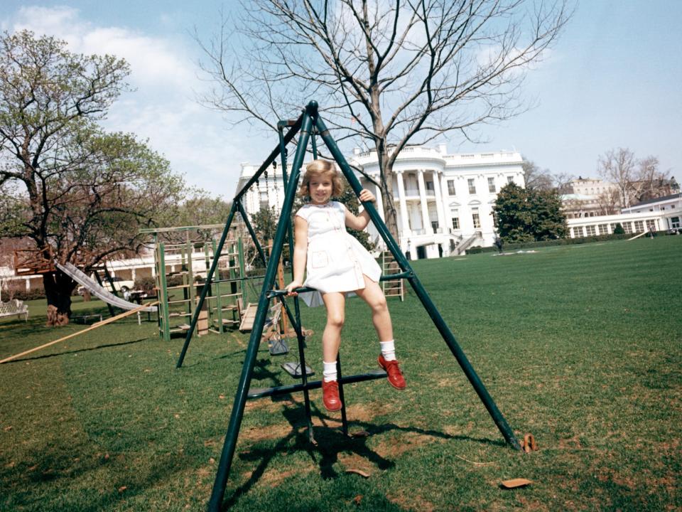 Caroline Kennedy, wearing red shoes, perches on a swing set on the White House lawn