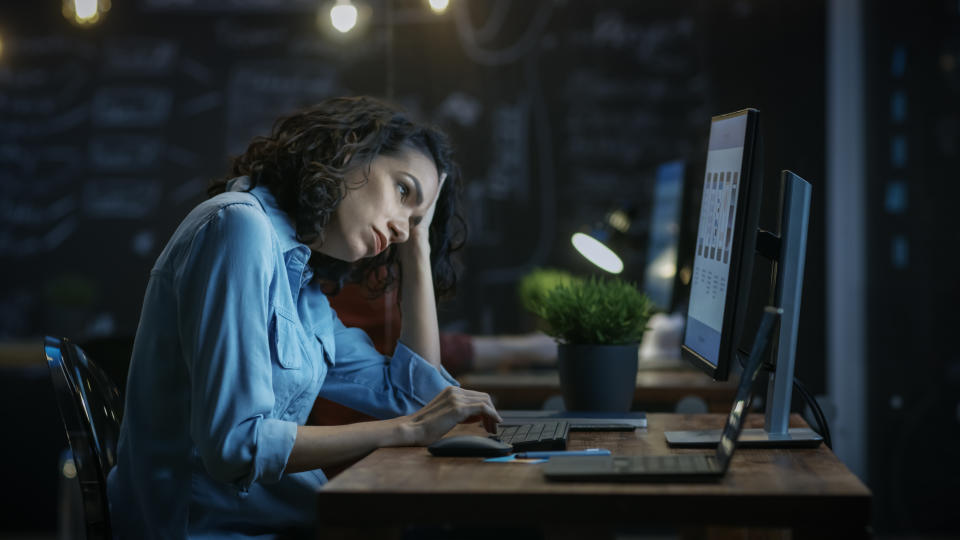 Tired, Overworked Female Financier Holds Her Head in Hands while Working on a Personal Computer. In the Background Creative Office.