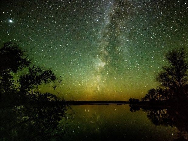 A photography of the Merritt Reservoir at night, with a clear view of the stars. Framed by trees on either side.