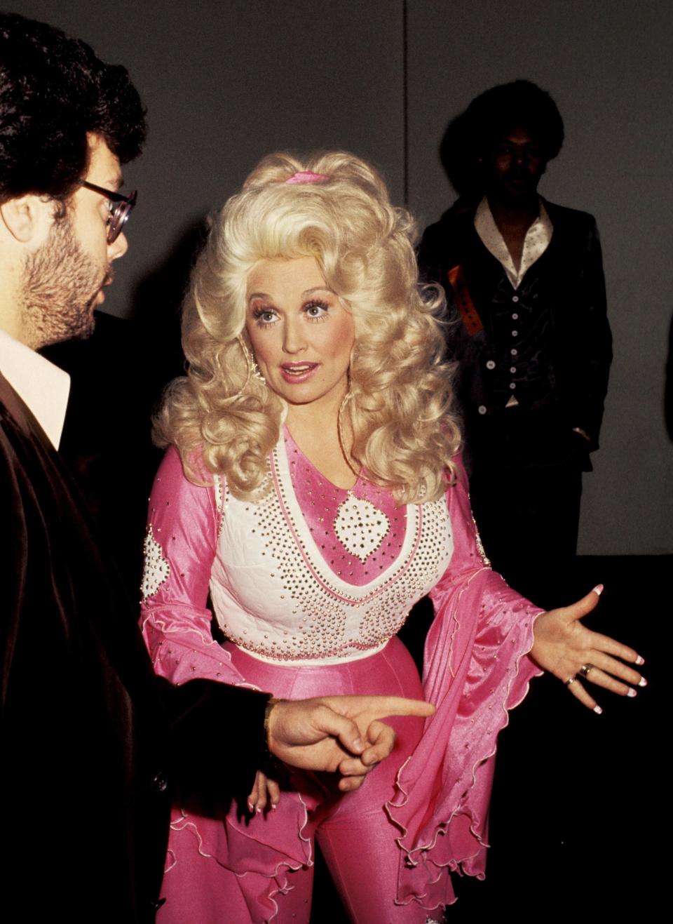 Dolly Parton in a bright pink and white blouse, vest, and pant ensemble.