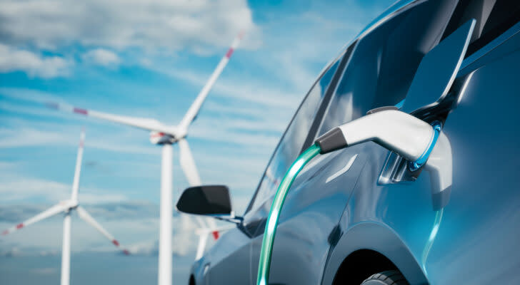 Photo of steel blue electric car being charged with wind silos and blue sky in the background