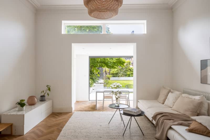 Neutral living room with airy doorways and windows with views of dining room and outdoors, accent tables, neutral rug and couch with neutral pillows and blanket, and wicker lamp.