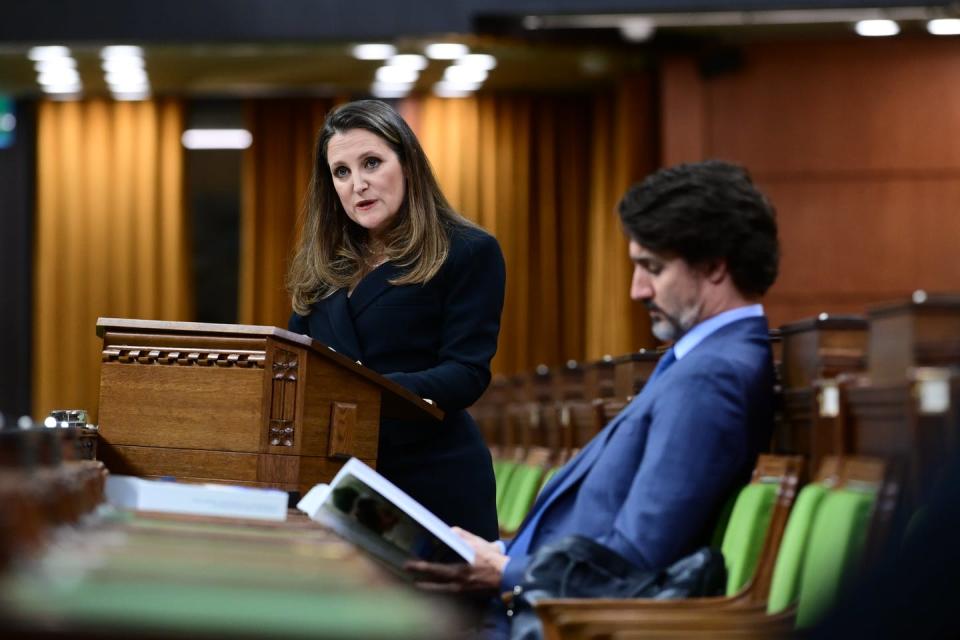 Chrystia Freeland stands at a podium to read the federal budget while Justin Trudeau leans back in his seat reading the budget document.