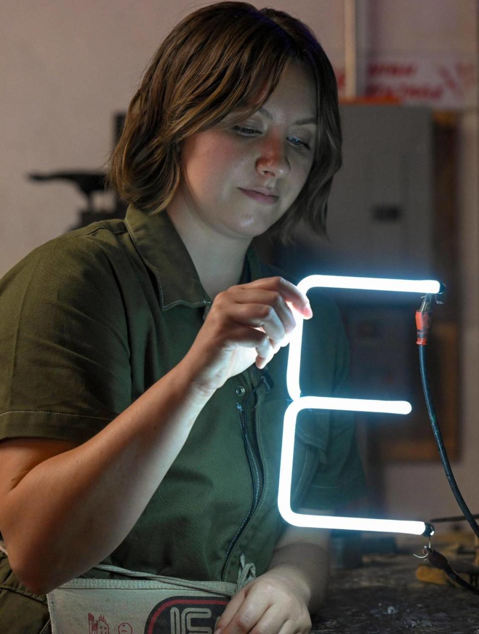 Neon tube bender Olivia Shelton, who works with her father, Randy Steinmetz, and brother, Dylan Steinmetz, checks the illumination of an “E” created at Element Ten, the family’s neon art and sign business.