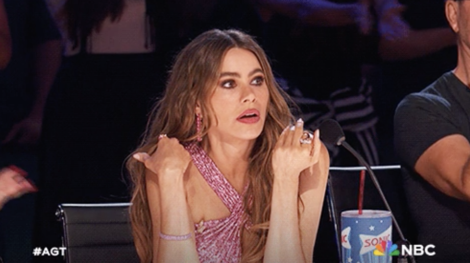 Sofía Vergara, on the judging panel of America's Got Talent, wearing a stylish one-shoulder dress, gestures with both hands while speaking into a microphone