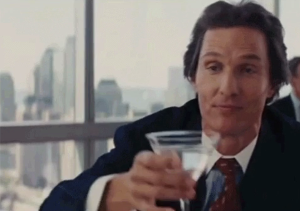 Matthew McConaughey in business attire making a toast with a drink, looking confident