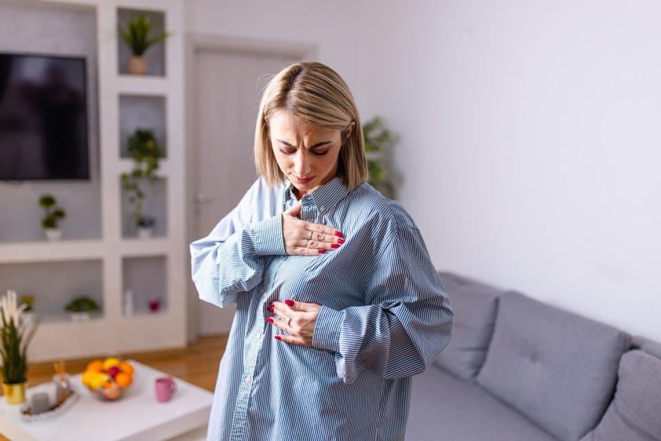 a young woman in casual clothes is standing in her living room and checking her breasts for possible lumps