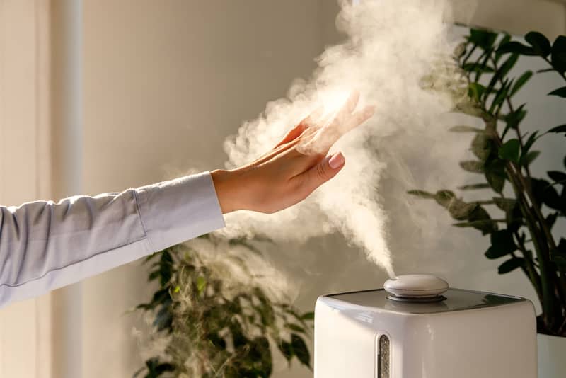 Woman holds hand over steam aroma oil diffuser on the table at home, steam from humidifier.