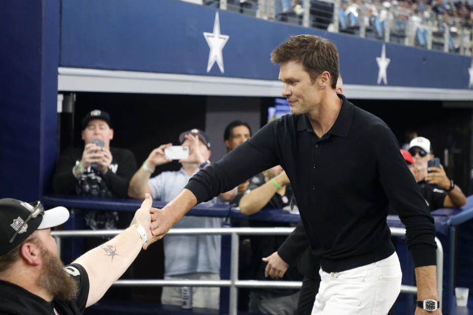 Former quarterback Tom Brady, right, greets fans as he walks off the field after players warm up for a preseason NFL football game between the Las Vegas Raiders and the Dallas Cowboys in Arlington, Texas, Saturday, Aug. 26, 2023. (AP Photo/Michael Ainsworth)