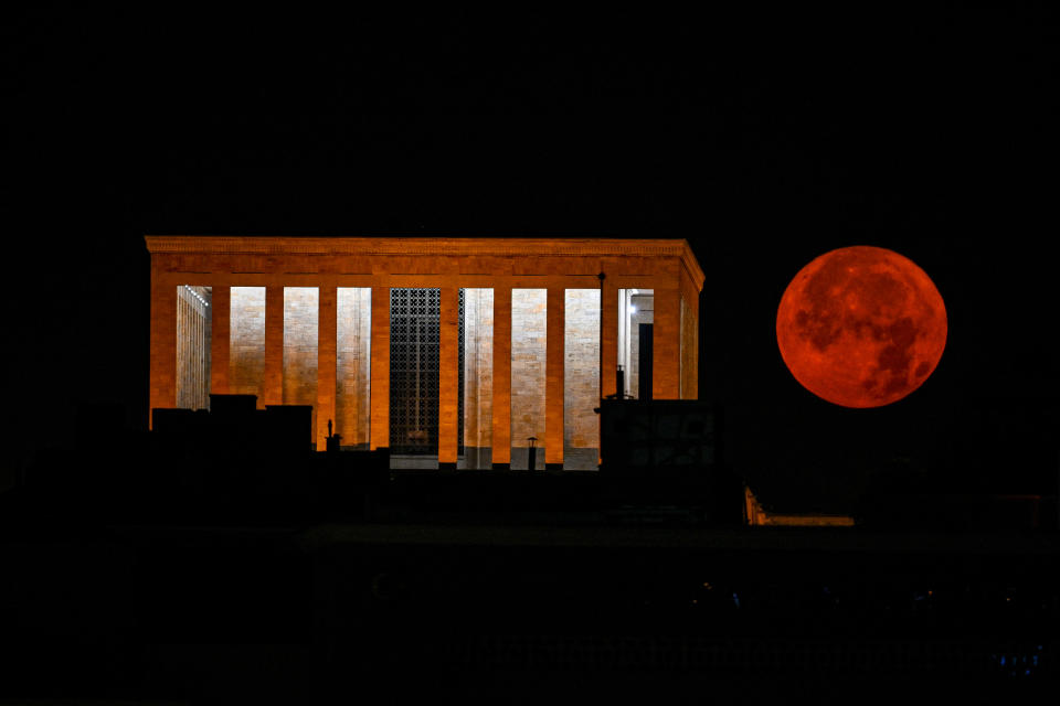 A large illuminated building on the left with eight large columns, next to a large blood red full moon
