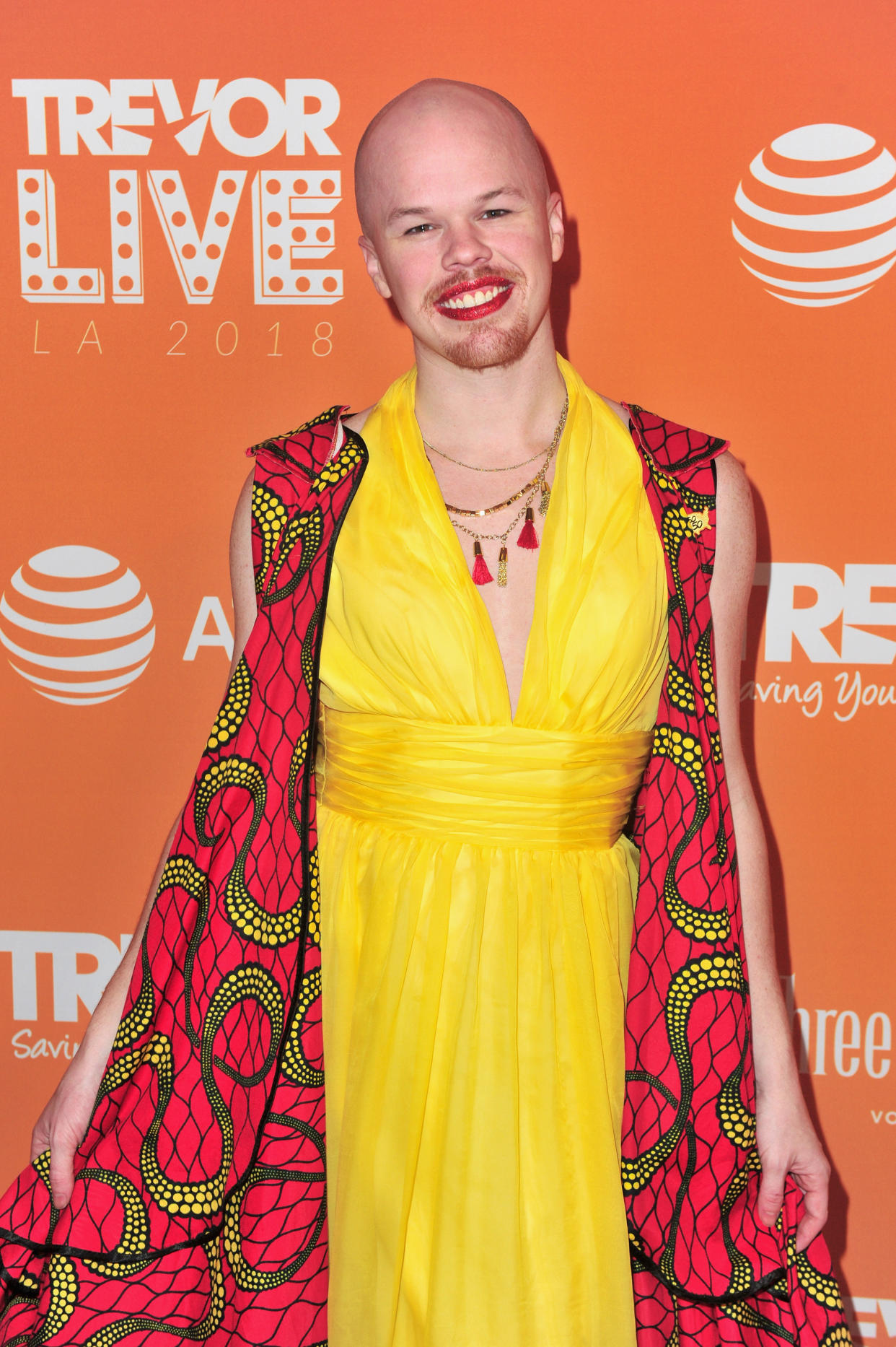 Sam Brinton wears a yellow dress and a red and gold print vest in front of a background that reads: Trevor Live.