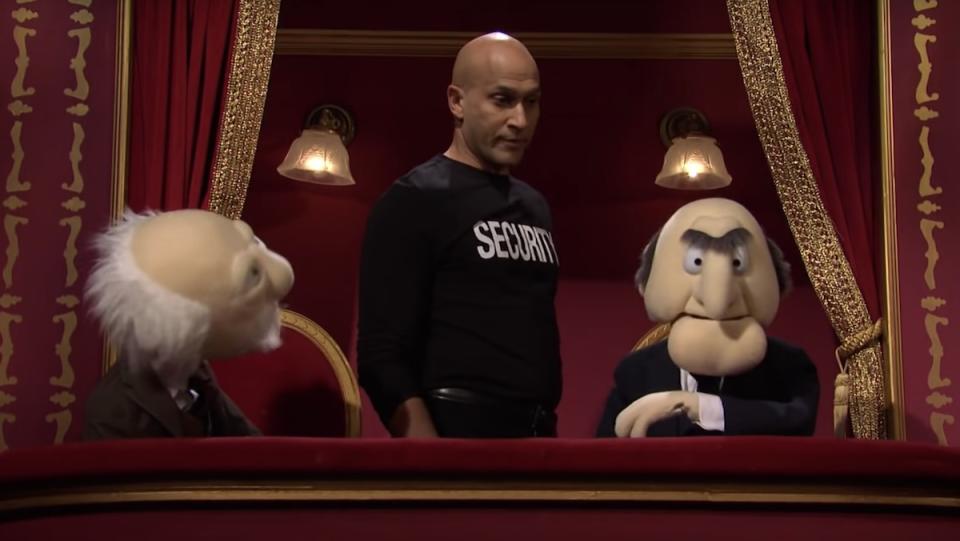 Keegan-Michael Key in a black security t-shirt stands behind Waldorf and Statler in their balcony box