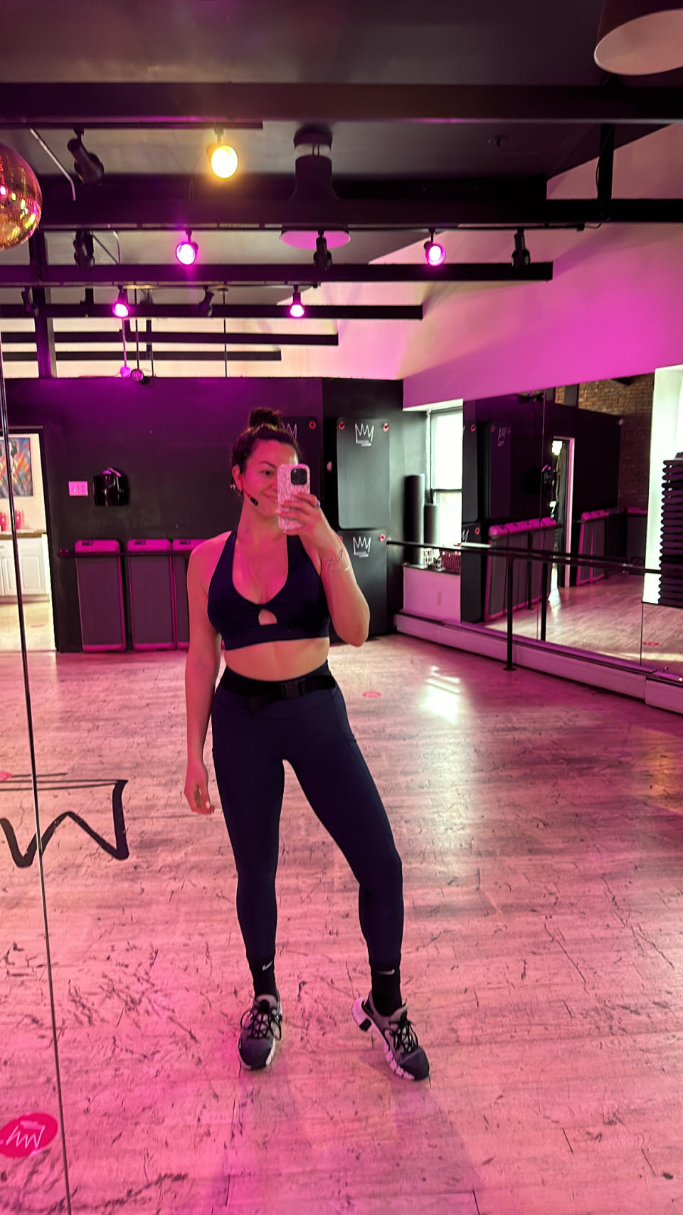 Person takes a mirror selfie in a gym studio. They wear a black sports bra and leggings with sneakers. Bright lights visible overhead