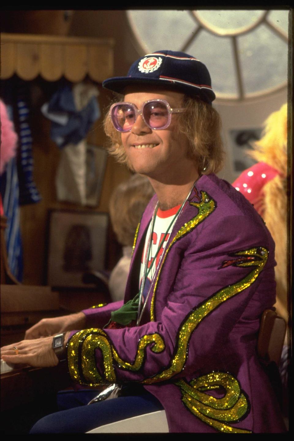 Elton John wears a vibrant jacket decorated with sparkling designs, a hat with a badge, and oversized glasses while sitting at a piano
