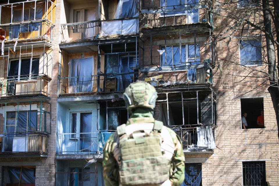 The back of a figure in a green camouflage jacket and helmet looking at a damaged apartment building