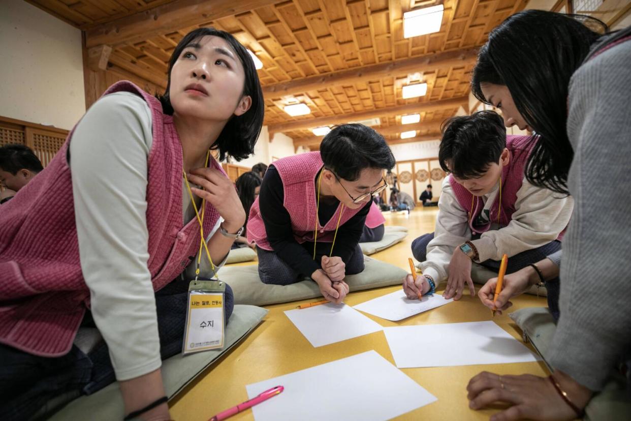 A woman in a pink vest looks to the left as three other people hold pencils near sheets of paper while seated on the floor