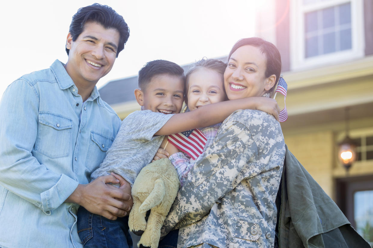 Smiling family look at the camera while embracing. The mom has returned from military assignment. The little girl is holding a small US flag.