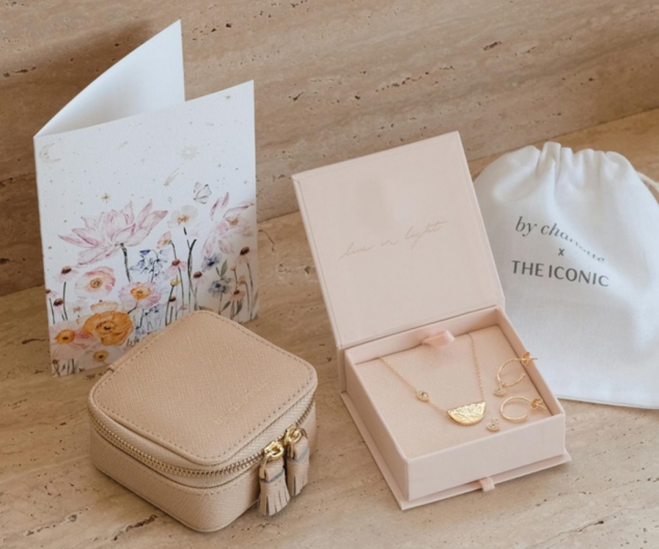 A beige mini leather square jewellery bag sits on beige stone with a pale pink jewellery cardboard box sits next to it on the right with the lid open showing a gold necklace and small gold hoop earrings. A floral card stands behind on the left with a small white draw string linen bag on the right.