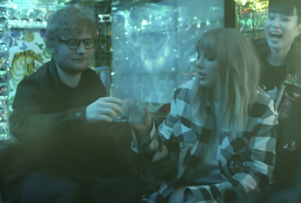 Ed Sheeran and Taylor Swift sitting together, wearing casual clothes, with a backdrop featuring blurred lights